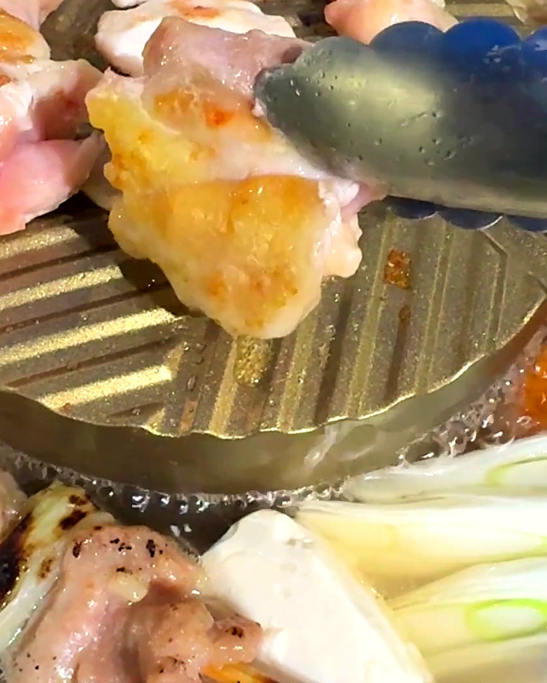 焼けた鶏肉をトングで持ち上げたところ。下では焼鳥串や豆腐、ねぎがスープでぐつぐつ煮えている